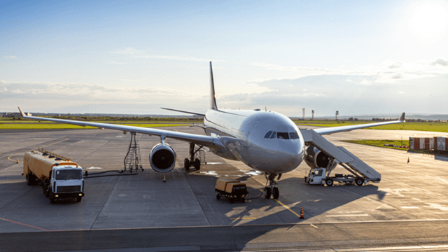 Commercial airplane parked at airport gate with boarding stairs, fuel truck, and ground support vehicles during sunset.